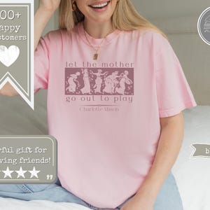 May include: A light pink short-sleeved t-shirt featuring a dark pink rectangular graphic with a line of figures and the text "let the mother go out to play" followed by "Charlotte Mason" in a serif font centred on the chest.