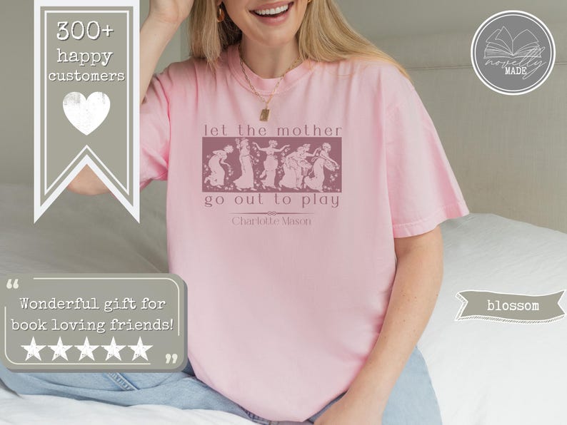 May include: A light pink short-sleeved t-shirt featuring a dark pink rectangular graphic with a line of figures and the text "let the mother go out to play" followed by "Charlotte Mason" in a serif font centred on the chest.