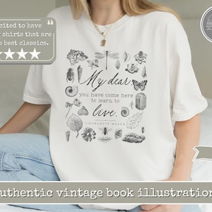 Pu&ograve; includere: Una t-shirt bianca con una stampa grafica nera di vari elementi naturali, tra cui insetti, conchiglie e foglie che circondano il testo: My dear you have come here to learn to live. Charlotte Mason. Il design &egrave; centrato sul petto.