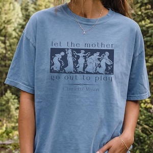 May include: A dusty blue short-sleeve t-shirt featuring a dark blue rectangular graphic with a classical illustration of five figures. The text reads: let the mother go out to play, followed by a decorative line and the name Charlotte Mason.