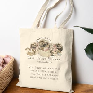 May include: Cream-colored tote bag featuring an illustration of three hedgehogs from "The Tale of Mrs. Tiggy-Winkle" by Beatrix Potter. The text on the bag includes the title and a quote from the story. The bag has two beige handles.