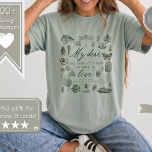 Pu&ograve; includere: Una t-shirt a maniche corte color verde salvia con una stampa grafica verde scuro di varie illustrazioni botaniche e di insetti che circondano il testo: My dear, you have come here to learn to live. Charlotte Mason &egrave; stampato sotto la citazione.
