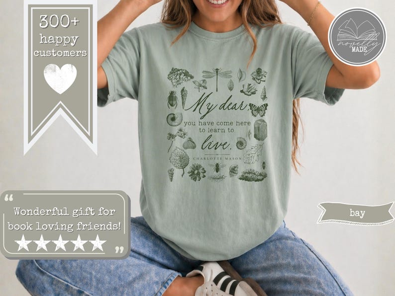 Pu&ograve; includere: Una t-shirt a maniche corte color verde salvia con una stampa grafica verde scuro di varie illustrazioni botaniche e di insetti che circondano il testo: My dear, you have come here to learn to live. Charlotte Mason &egrave; stampato sotto la citazione.