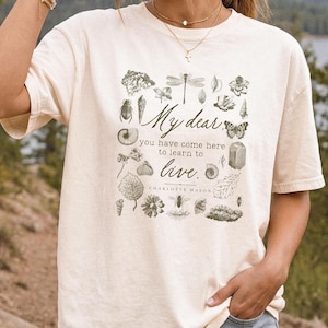 Pu&ograve; includere: Una t-shirt a maniche corte color crema con una stampa grafica verde scuro di varie illustrazioni botaniche e di insetti che circondano il testo: My dear, you have come here to learn to live. CHARLOTTE MASON.