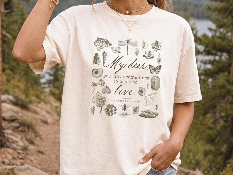 Pu&ograve; includere: Una t-shirt a maniche corte color crema con una stampa grafica verde scuro di varie illustrazioni botaniche e di insetti che circondano il testo: My dear, you have come here to learn to live. CHARLOTTE MASON.