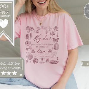 Pu&ograve; includere: Una t-shirt a maniche corte rosa chiaro con una stampa grafica marrone scuro di vari elementi naturali, tra cui insetti, conchiglie e foglie. Il testo recita: My dear, you have come here to learn to live. Charlotte Mason.