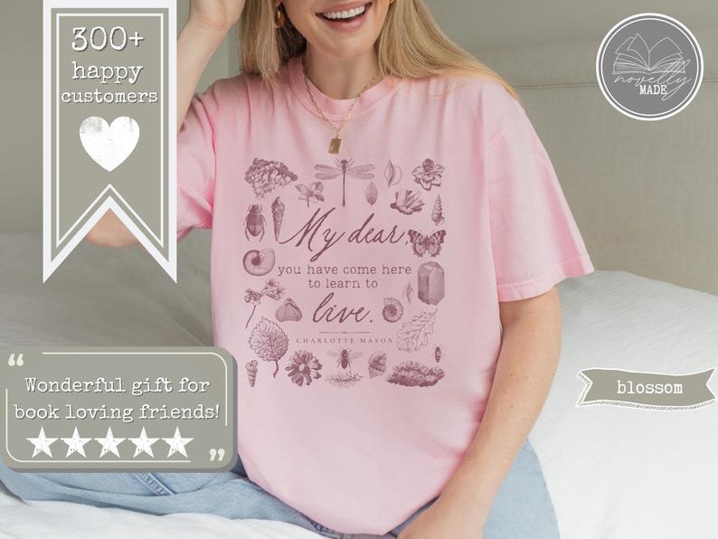 Pu&ograve; includere: Una t-shirt a maniche corte rosa chiaro con una stampa grafica marrone scuro di vari elementi naturali, tra cui insetti, conchiglie e foglie. Il testo recita: My dear, you have come here to learn to live. Charlotte Mason.