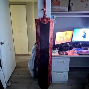 May include: A large, handmade sword prop with a dark blade and a red hilt. The sword is mounted on a wall, showcasing its size and detailed craftsmanship. The blade appears to be approximately 60 inches long.