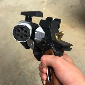 May include: A futuristic prop pistol featuring a black frame, a brown grip, and a silver-colored six-chambered cylinder. The device includes a top-mounted rail, a gold-colored hammer, and a silver accent piece at the base of the handle.