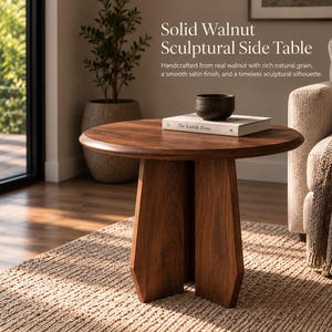 May include: A round side table made of solid walnut wood with a smooth finish and a sculptural base. The table features a circular top and a central support structure. A dark wooden bowl and a book titled The Kinfolk Home sit on top.