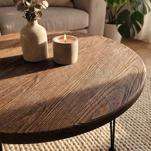 Może przedstawiać: Okrągły drewniany stolik kawowy z postarzanym wykończeniem. Na stole stoi zapalona świeca w betonowym świeczniku i ceramiczny wazon z suszonymi kwiatami. Stół ma czarne metalowe nogi.