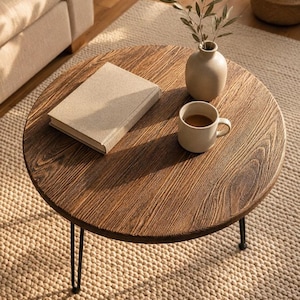 Peut inclure: Une table basse ronde en bois avec une finition grain de bois brun foncé et trois pieds en épingle en métal noir. Sur le plateau se trouvent un livre beige fermé, un petit vase en céramique beige avec un brin feuillu et une tasse beige.