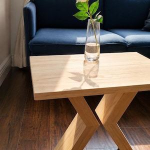 Peut inclure: Une table d'appoint en bois clair avec un plateau rectangulaire et une base géométrique en forme de X. Un vase en verre transparent contenant une rose blanche unique avec des feuilles vertes est posé sur la surface, projetant une ombre.