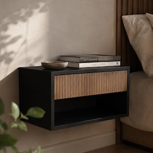 May include: A wall-mounted floating nightstand featuring a matte black rectangular frame and a single drawer front with vertical wood slats. A small stone bowl and two books titled Axel Vervoordt Wabi Inspirations and This Is Home sit on top.