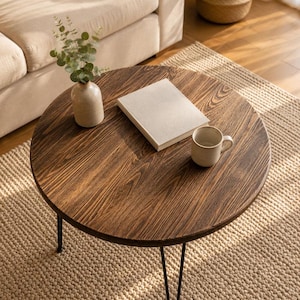 Puede incluir: Mesa de centro redonda de madera oscura con patas de horquilla negras. Un libro beige, una taza de cerámica y un pequeño jarrón con vegetación están sobre la mesa. La mesa está sobre una alfombra beige.