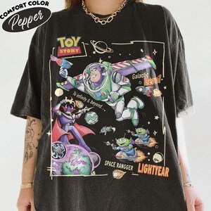 May include: A black t-shirt featuring a Toy Story graphic with Buzz Lightyear, Zurg, and aliens in space. The print includes the text Toy Story, To infinity & beyond, Galactic Hero, SPACE RANGGER, and LIGHTYEAR. The shirt has a crew neck.