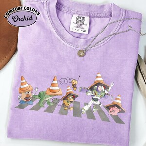 K&ouml;nnte beinhalten: Ein lila Comfort Colors T-Shirt mit einer Grafik von Toy Story-Charakteren, die einen Zebrastreifen &uuml;berqueren, jeder mit einem Verkehrskegel-Hut. Das Shirt hat ein kleines Comfort Colors-Etikett und eine silberne Halskette.