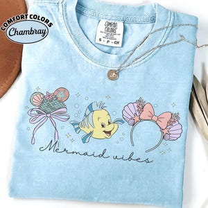May include: Light blue t-shirt with the words "Mermaid Vibes" and a graphic of a yellow fish and mermaid-themed accessories. The shirt has a crew neck and short sleeves. A silver necklace with a pendant is on the shirt.