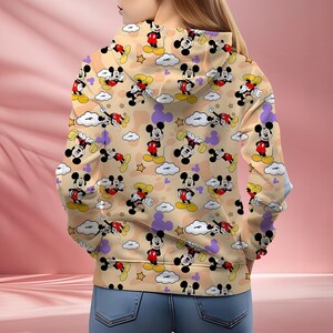 K&ouml;nnte beinhalten: Ein beigefarbener Kapuzenpullover mit einem sich wiederholenden Muster von Mickey Mouse-Charakteren in verschiedenen Posen. Das Design umfasst Wolken, Sterne und lila Mickey Mouse-Kopfformen. Der Kapuzenpullover wird von einer Person getragen, die blaue Jeans tr&auml;gt.