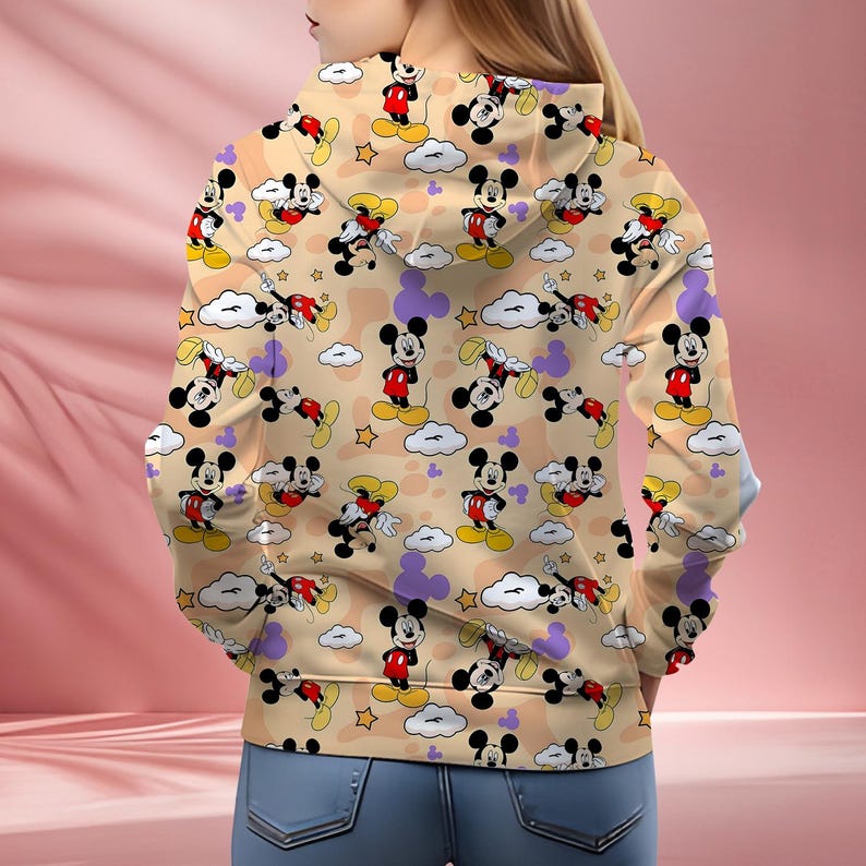 K&ouml;nnte beinhalten: Ein beigefarbener Kapuzenpullover mit einem sich wiederholenden Muster von Mickey Mouse-Charakteren in verschiedenen Posen. Das Design umfasst Wolken, Sterne und lila Mickey Mouse-Kopfformen. Der Kapuzenpullover wird von einer Person getragen, die blaue Jeans tr&auml;gt.