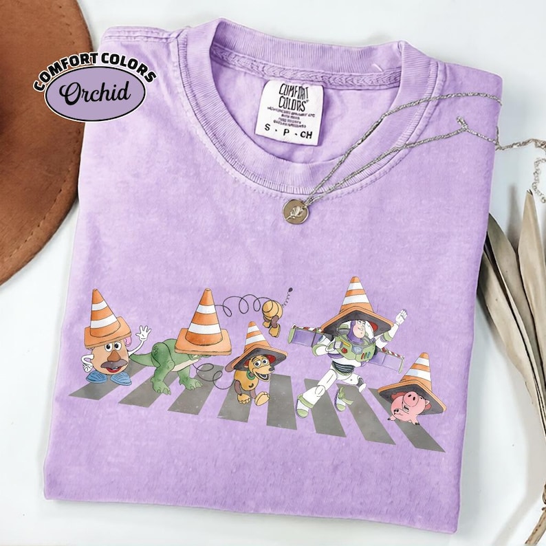K&ouml;nnte beinhalten: Ein lila Comfort Colors T-Shirt mit einer Grafik von Toy Story-Charakteren, die einen Zebrastreifen &uuml;berqueren, jeder mit einem Verkehrskegel-Hut. Das Shirt hat ein kleines Comfort Colors-Etikett und eine silberne Halskette.