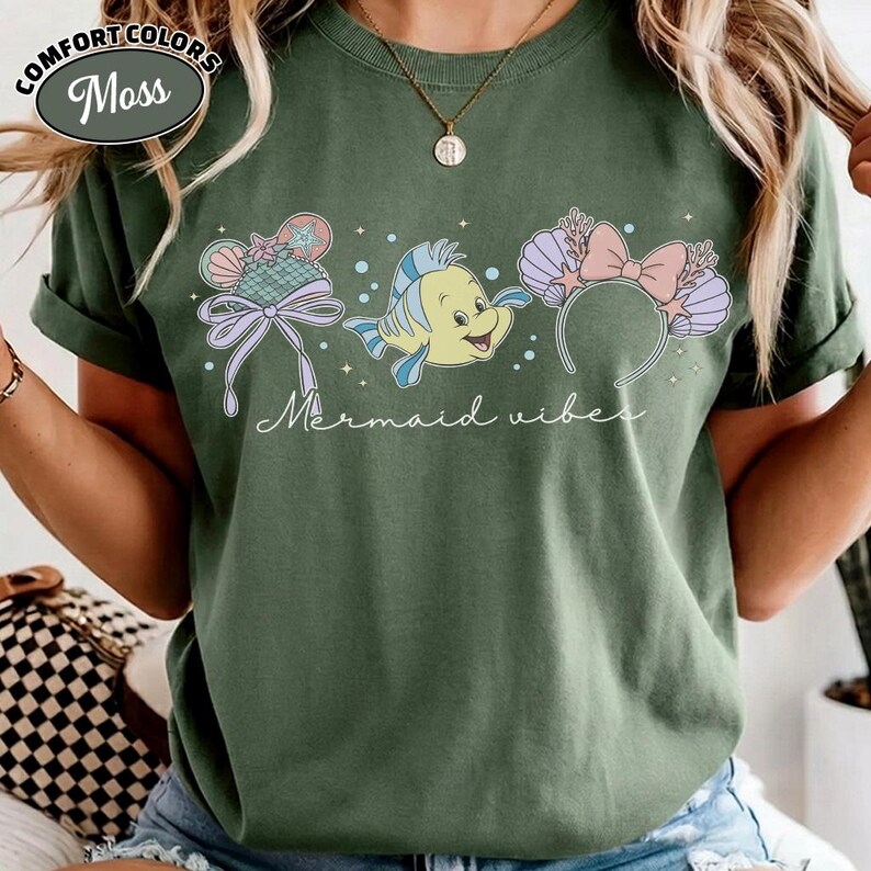 May include: Green Comfort Colors t-shirt with a graphic of a yellow fish and mermaid-themed headbands. The text "Mermaid vibes" is written in cursive. The shirt has rolled-up sleeves and a round neckline.