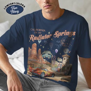 Puede incluir: Camiseta azul marino con un gr&aacute;fico de Radiator Springs, California, con un coche de carreras, un paisaje des&eacute;rtico y el texto "Radiator Springs Est. 2012". La camiseta es de Comfort Colors.