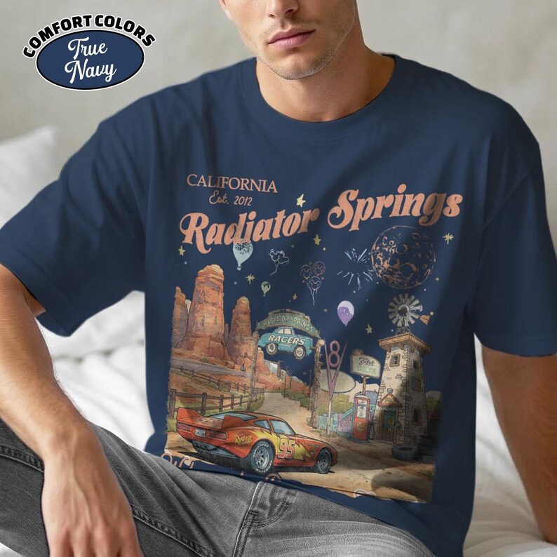 Puede incluir: Camiseta azul marino con un gr&aacute;fico de Radiator Springs, California, con un coche de carreras, un paisaje des&eacute;rtico y el texto "Radiator Springs Est. 2012". La camiseta es de Comfort Colors.