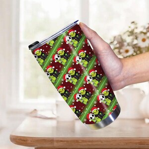 May include: A stainless steel tumbler with a repeating pattern of a green cartoon character wearing a Santa hat and a black and white character wearing a Santa hat. The background is red with green stripes and snowflakes.