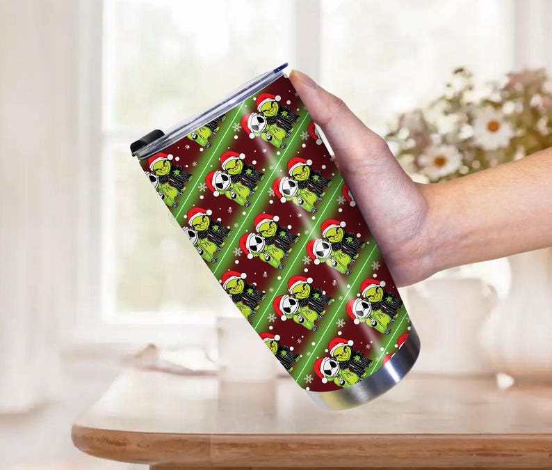 May include: A stainless steel tumbler with a repeating pattern of a green cartoon character wearing a Santa hat and a black and white character wearing a Santa hat. The background is red with green stripes and snowflakes.