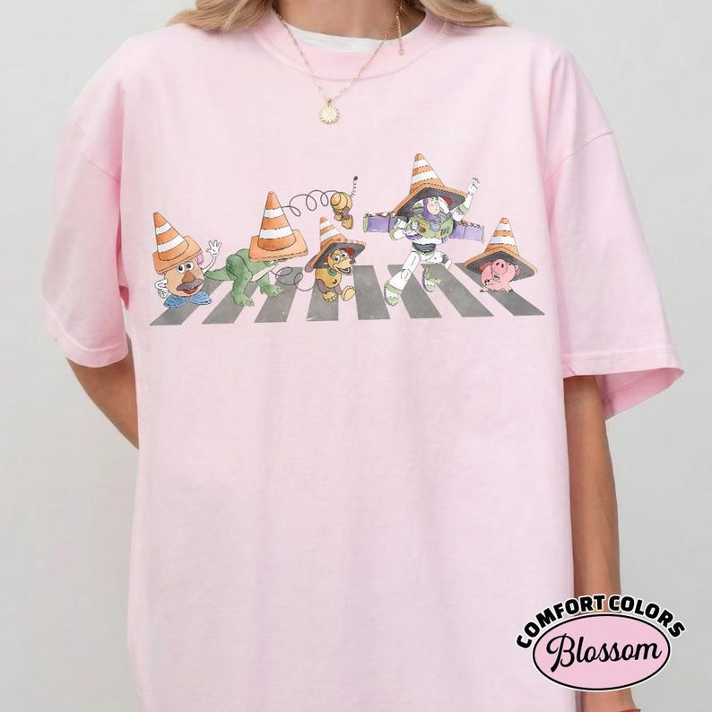 K&ouml;nnte beinhalten: Hellrosa T-Shirt mit einer Cartoon-Illustration von Toy Story-Charakteren, die eine Stra&szlig;e &uuml;berqueren, jeder mit einem Verkehrskegel-Hut. Das Design befindet sich auf einem Comfort Colors Blossom Shirt.