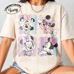 May include: Ivory Comfort Colors t-shirt featuring four panels of Minnie Mouse in different poses. Minnie is wearing a pink bow and various outfits. The shirt has the text "Comfort Colors Ivory" in a circular design.