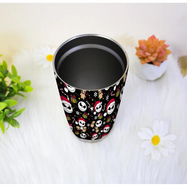 May include: A stainless steel tumbler with a black background featuring a pattern of Jack Skellington in Santa hats, gifts, snowflakes, and candy canes. The tumbler has a silver interior and is shown on a white, fluffy surface.