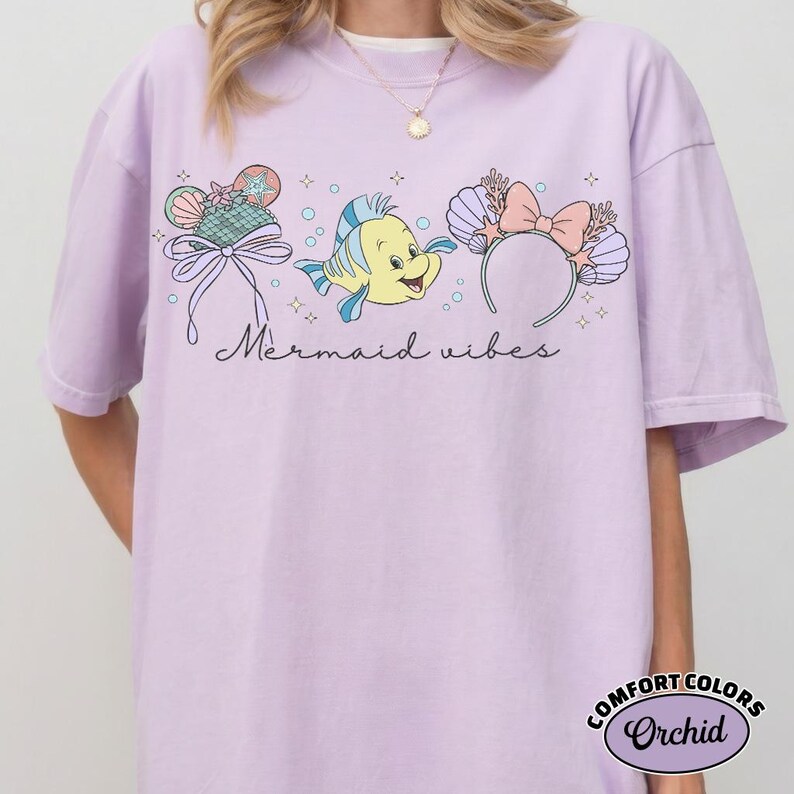 May include: A light purple t-shirt with the words "Mermaid vibes" in cursive. The shirt features three cartoon graphics: a seashell-shaped hat, a yellow fish, and a seashell headband with a pink bow. The shirt is labeled "Comfort Colors Orchid."