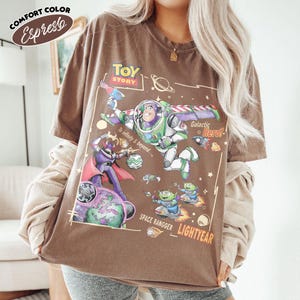 May include: An espresso brown short-sleeve t-shirt featuring a graphic print of Buzz Lightyear, Zurg, and aliens from Toy Story. The design includes the text "Toy Story," "To infinity & beyond," "Galactic Hero," and "Space Rangger Lightyear."