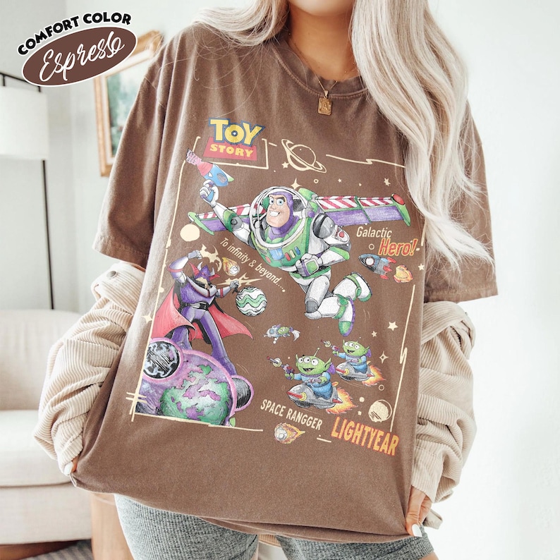 May include: An espresso brown short-sleeve t-shirt featuring a graphic print of Buzz Lightyear, Zurg, and aliens from Toy Story. The design includes the text "Toy Story," "To infinity & beyond," "Galactic Hero," and "Space Rangger Lightyear."