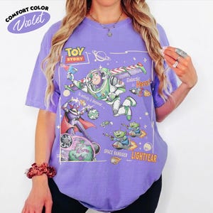 May include: A violet short-sleeve t-shirt featuring a Toy Story graphic print. The design includes Buzz Lightyear, Zurg, and green aliens with the text Toy Story, Galactic Hero, To infinity & beyond, Space Rangger, and Lightyear in a space theme.