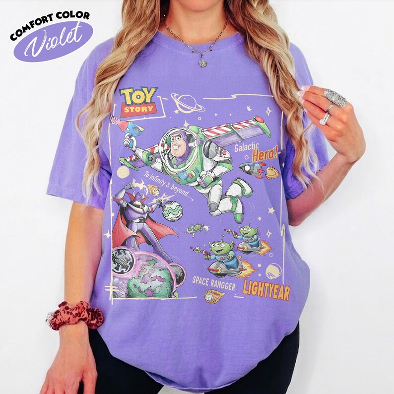 May include: A violet short-sleeve t-shirt featuring a Toy Story graphic print. The design includes Buzz Lightyear, Zurg, and green aliens with the text Toy Story, Galactic Hero, To infinity & beyond, Space Rangger, and Lightyear in a space theme.