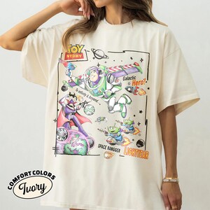 May include: An ivory t-shirt featuring a Toy Story graphic with Buzz Lightyear, Zurg, and green aliens. The design includes the text TOY STORY, Galactic Hero!, To infinity & beyond..., SPACE RANGGER, and LIGHTYEAR in a colorful, hand-drawn style.