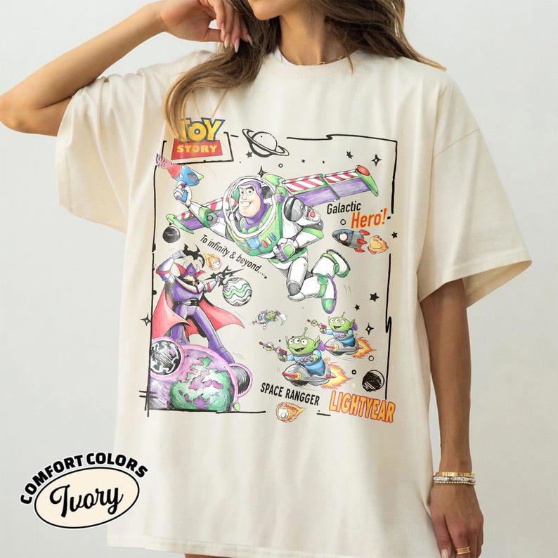 May include: An ivory t-shirt featuring a Toy Story graphic with Buzz Lightyear, Zurg, and green aliens. The design includes the text TOY STORY, Galactic Hero!, To infinity & beyond..., SPACE RANGGER, and LIGHTYEAR in a colorful, hand-drawn style.
