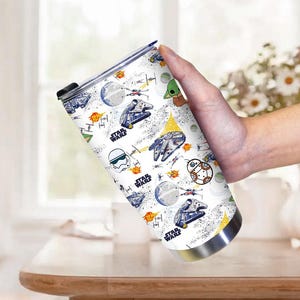 Op de afbeelding: Een witte reismok met een Star Wars-thema. Het ontwerp bevat illustraties van ruimteschepen, personages zoals Baby Yoda en het Star Wars-logo. De mok heeft een zilveren basis en een zwart deksel.