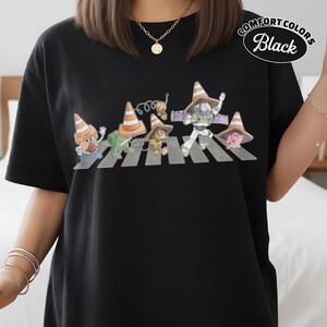 K&ouml;nnte beinhalten: Schwarzes T-Shirt mit einer Parodie des Abbey Road-Albumcovers mit Toy Story-Charakteren, die Verkehrskegel tragen. Der Text "Comfort Colors Black" ist oben rechts auf dem Shirt aufgedruckt.