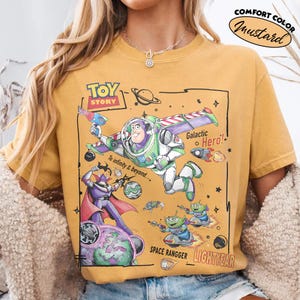 May include: A mustard yellow t-shirt featuring a graphic print of Buzz Lightyear, Zurg, and aliens from Toy Story. The design includes the text "Toy Story", "Galactic Hero!", "To infinity & beyond....", "SPACE RANGGER", and "LIGHTYEAR".
