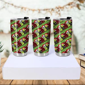 May include: Three stainless steel insulated tumblers with black lids. Each tumbler features a repeating pattern of green characters wearing Santa hats on a red background with green diagonal stripes and snowflakes.