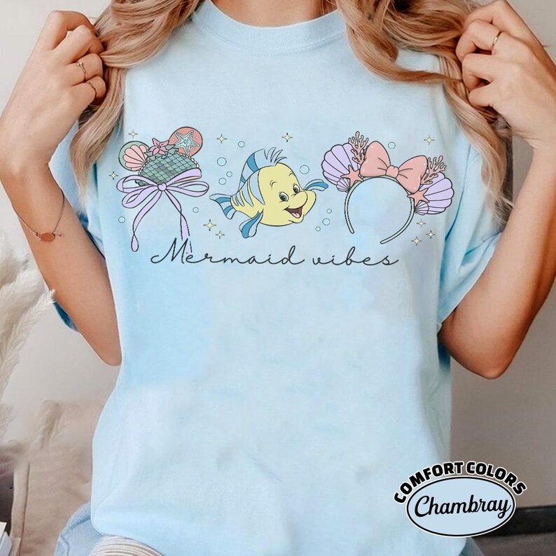 May include: Light blue t-shirt with a graphic featuring a cartoon fish, seashell headband, and a decorative bow. The text "Mermaid vibes" is written in cursive. The shirt is made of Comfort Colors Chambray material.