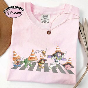 K&ouml;nnte beinhalten: Rosa Comfort Colors T-Shirt mit einem Aufdruck von Toy Story-Charakteren, die einen Zebrastreifen &uuml;berqueren, jeder mit einem Verkehrskegel-Hut. Der Text "Comfort Colors Blossom" ist ebenfalls abgebildet.