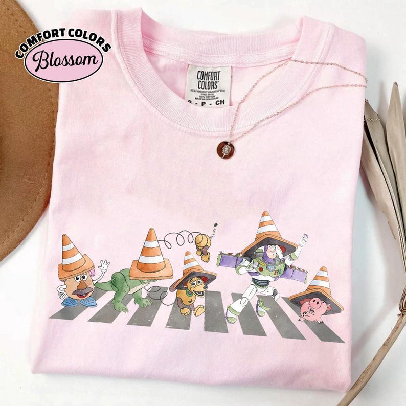 K&ouml;nnte beinhalten: Rosa Comfort Colors T-Shirt mit einem Aufdruck von Toy Story-Charakteren, die einen Zebrastreifen &uuml;berqueren, jeder mit einem Verkehrskegel-Hut. Der Text "Comfort Colors Blossom" ist ebenfalls abgebildet.