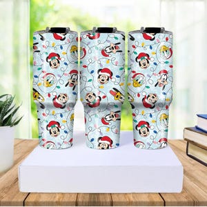 May include: Three stainless steel tumblers with a light blue background, featuring a repeating pattern of Mickey Mouse, Donald Duck, and Goofy in Santa hats, along with Christmas lights. The tumblers are displayed on a white surface.