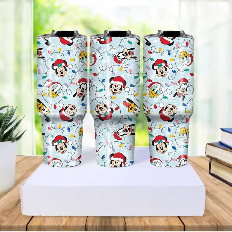 May include: Three stainless steel tumblers with a light blue background, featuring a repeating pattern of Mickey Mouse, Donald Duck, and Goofy in Santa hats, along with Christmas lights. The tumblers are displayed on a white surface.