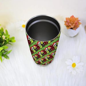 May include: A stainless steel tumbler with a wraparound pattern of green creatures wearing Santa hats on a red and green striped background. The tumbler has a black interior and a silver rim. The design is repeated diagonally.